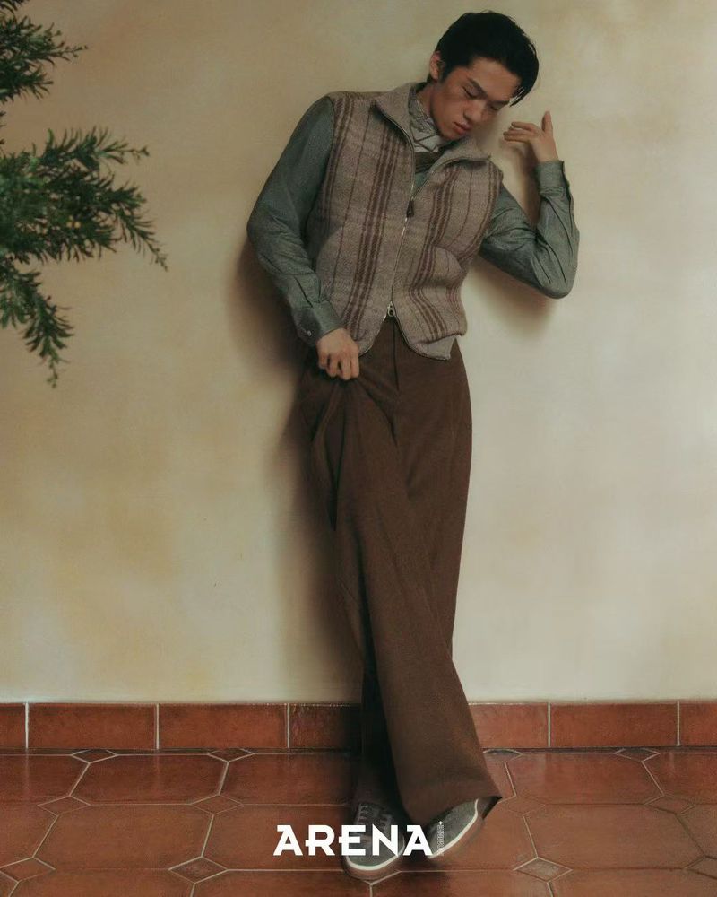 Male fashion model in vest and wide-legged trousers poses indoors on tiled floor, hand near head, in magazine editorial-style shot.