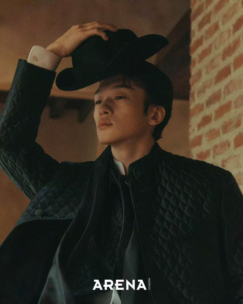 Man wearing a black quilted jacket touching his hat against a brick wall for Arena Homme Plus Korea