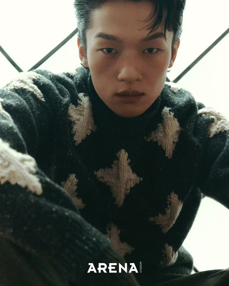 A young adult male poses indoors wearing a patterned sweater, creating a fashion-forward editorial portrait with neutral tones and soft lighting.