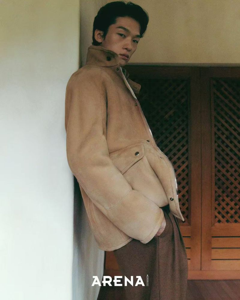 Model Jiaxuan Yang wearing a tan suede jacket and brown trousers, leaning against a wall next to a wooden screen.