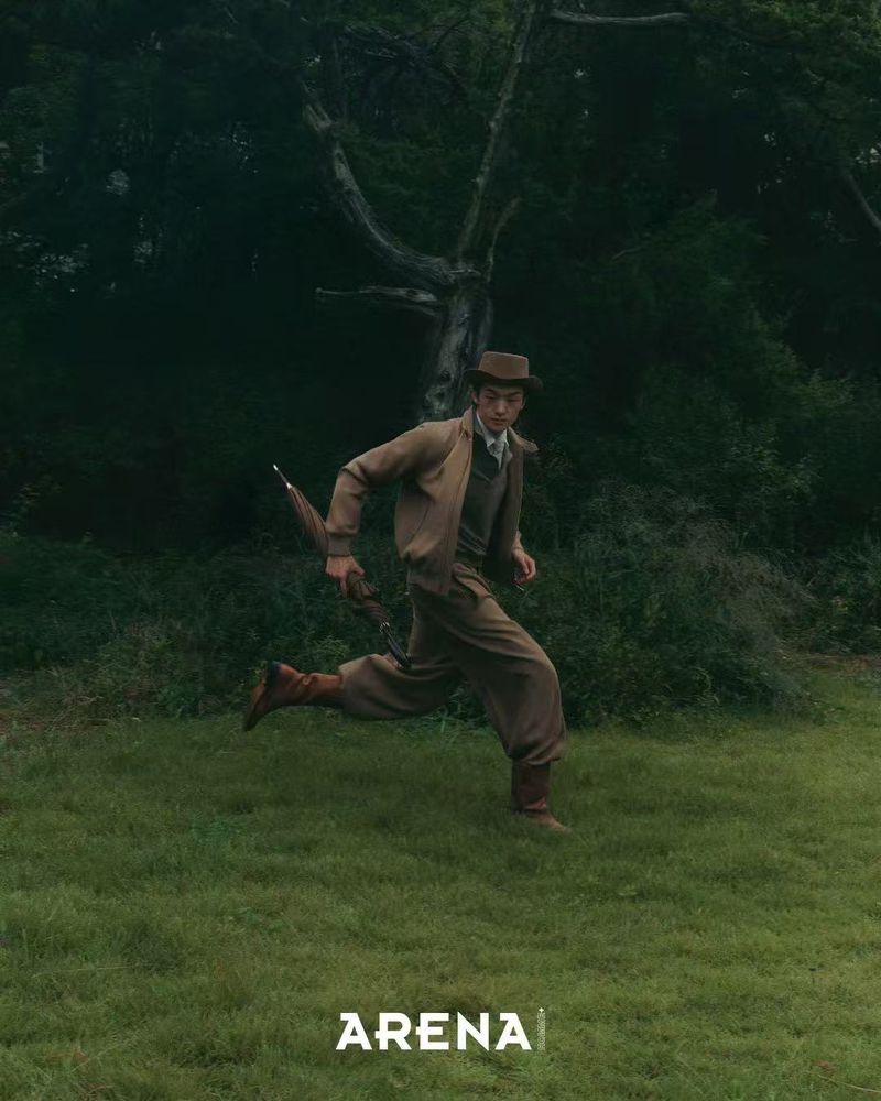 A man in vintage clothing runs across a green field, holding an umbrella against a dark forest backdrop, photographed for Arena magazine.