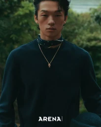 Close-up portrait of a man wearing a dark sweater, neck scarf, and gold necklace with lush green foliage blurred in the background.