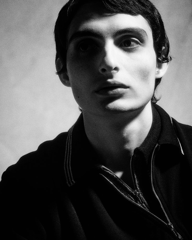 Portrait of a young adult male with dark hair looking off-camera in a high-contrast black and white studio lighting setup, wearing a dark zippered sweater.