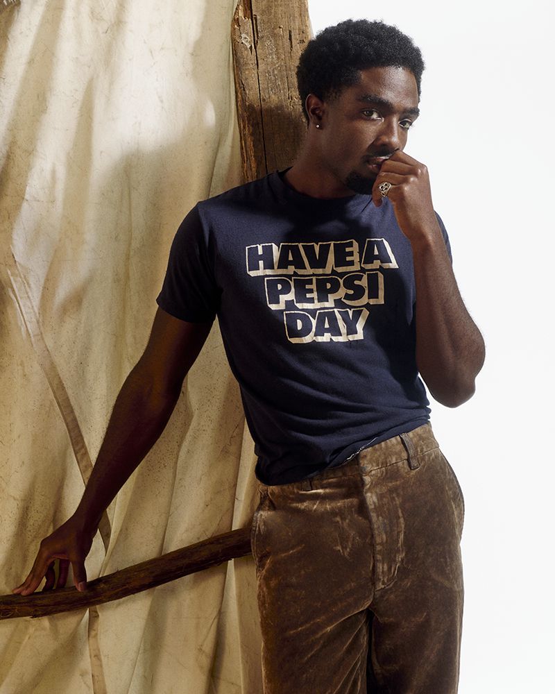 Young African-descent man in casual clothing poses with hand near face in a photo studio with soft lighting and neutral backdrop.