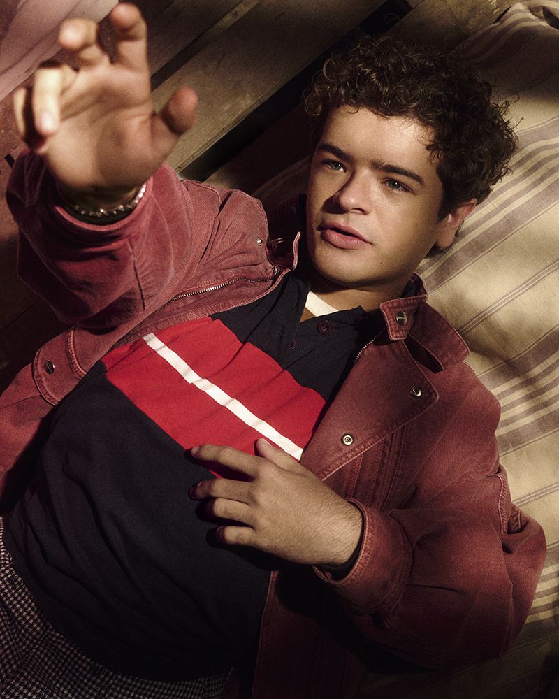 Young man lying on striped surface, wearing a red jacket and collared shirt, hand raised, looking at the camera in indoor setting.