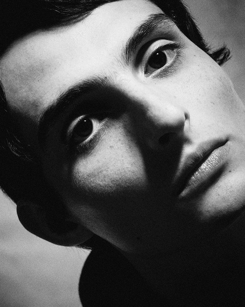 Close-up black and white portrait of a fair-skinned young adult male looking up at the camera with a neutral expression in a studio.