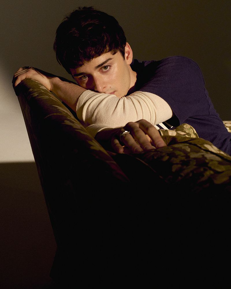 Portrait of a young adult male leaning on an upholstered object, with soft lighting highlighting his features and a dark background.