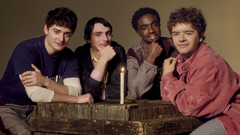 Four young men gather around a rustic wooden table illuminated by a single candle, each with unique attire and expressions.