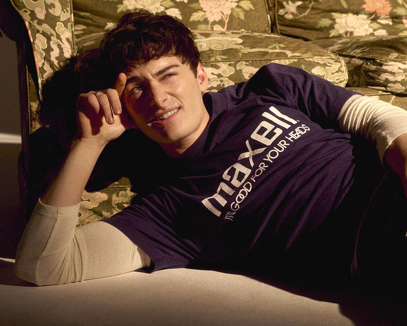 Young adult male lounging on a floral couch wearing a Maxell t-shirt smiles towards the camera in a warm-lit indoor setting.