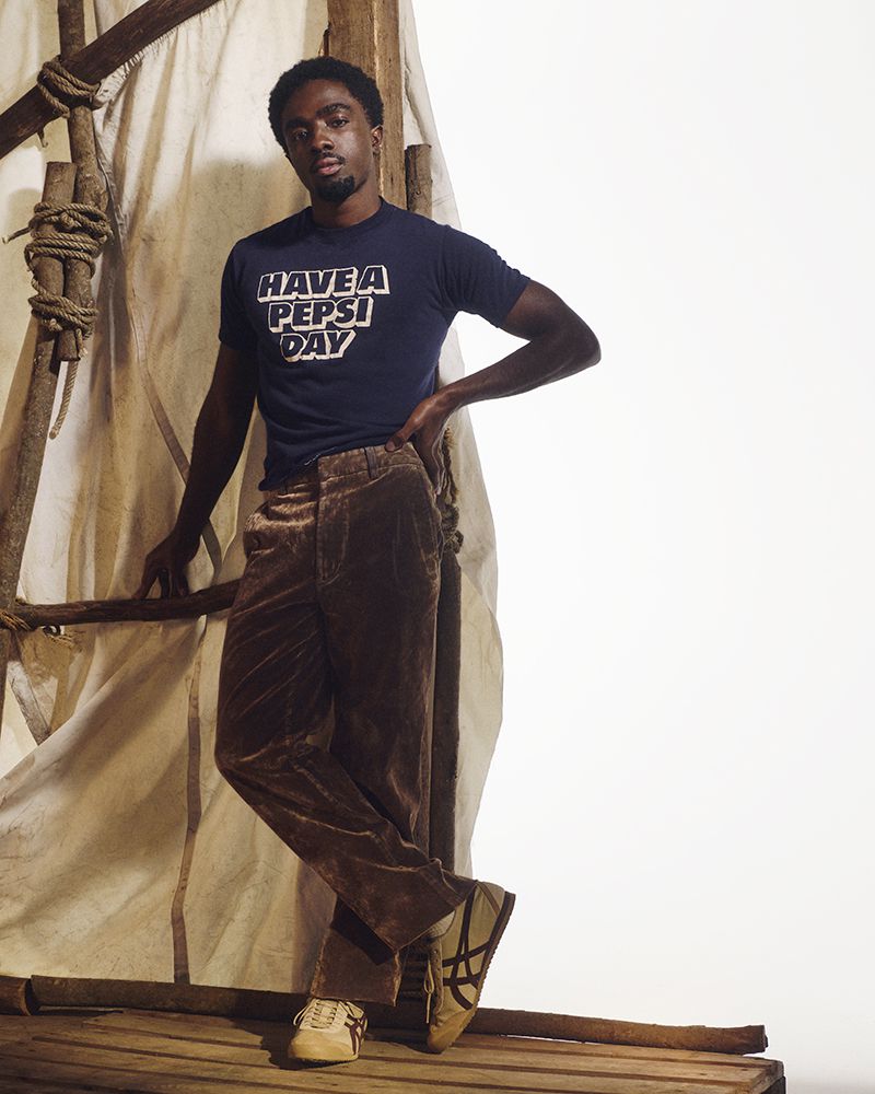 Adult male poses in t-shirt and brown velvet trousers, standing on rough wood planking, and set against a textured white backdrop.