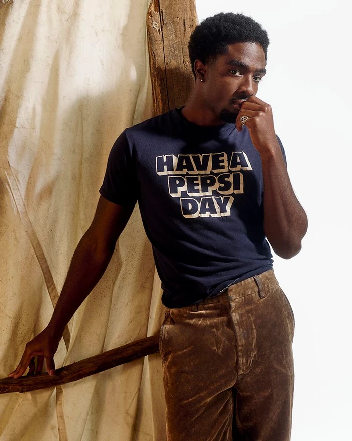 Caleb McLaughlin in a dark t-shirt and brown trousers leaning against a textured backdrop for British GQ.
