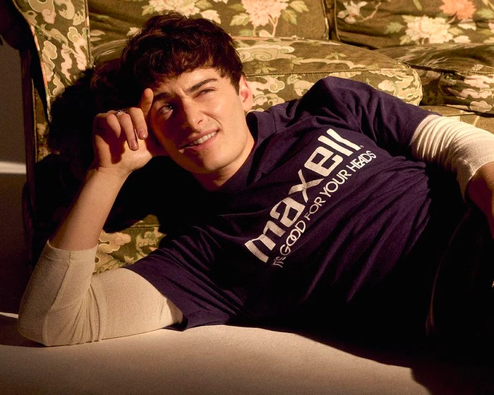 Noah Schnapp reclining on the floor against a vintage floral sofa wearing a navy graphic t-shirt over a white long-sleeve shirt.