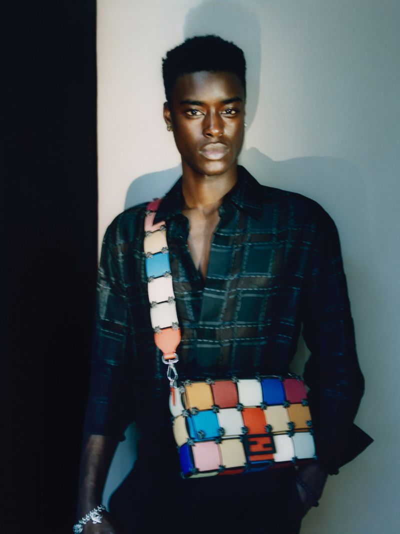 Young Black man in plaid shirt poses with colorful block-patterned crossbody bag in a fashion shot with neutral studio backdrop and dark accents.