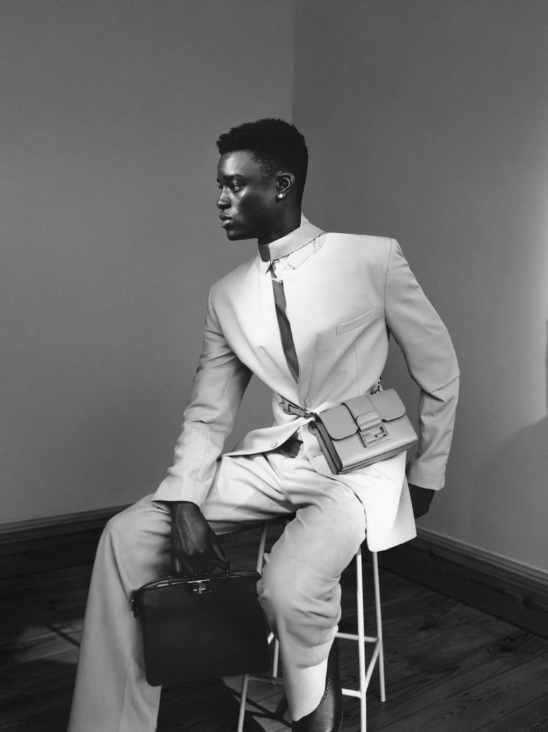 Black male model seated on a stool in a studio. He is wearing a light-colored suit, a dark tie, and carrying a black briefcase.