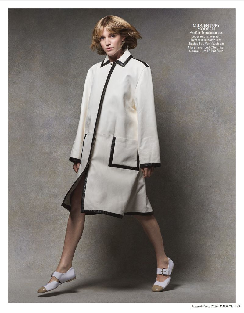 A woman poses in a white coat with black trim and two-tone Mary Jane shoes against a mottled grey backdrop in a fashion editorial.