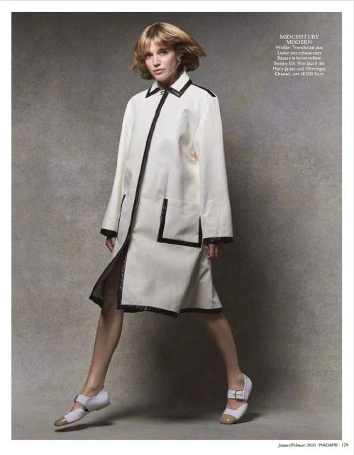A woman poses in a white coat with black trim and two-tone Mary Jane shoes against a mottled grey backdrop in a fashion editorial.