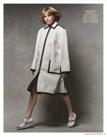A woman poses in a white coat with black trim and two-tone Mary Jane shoes against a mottled grey backdrop in a fashion editorial.