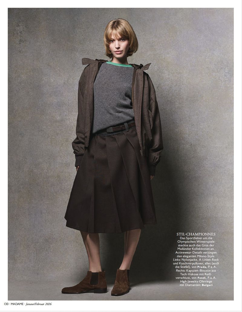 Fashion editorial shot: A young woman with short blonde hair poses in a grey sweater, brown jacket, brown skirt, and brown boots.