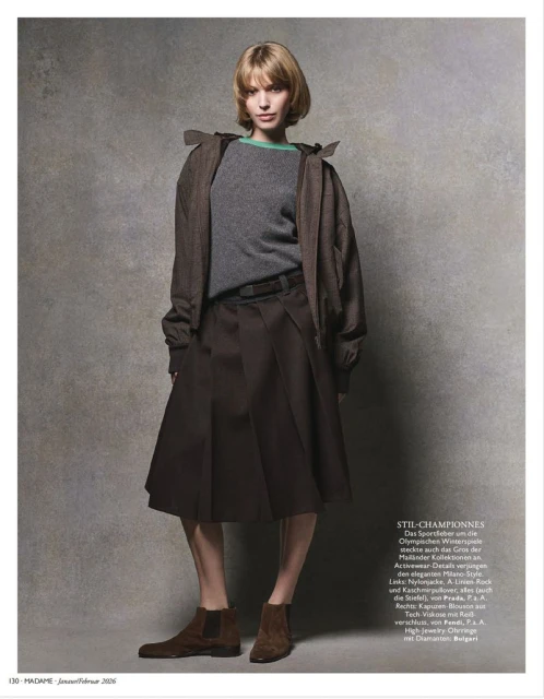 Fashion editorial shot: A young woman with short blonde hair poses in a grey sweater, brown jacket, brown skirt, and brown boots.