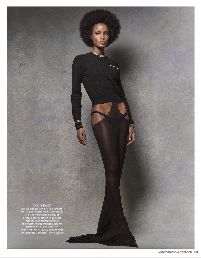 Model with afro hair wearing a black long-sleeve top and sheer long skirt against a mottled grey textured backdrop.