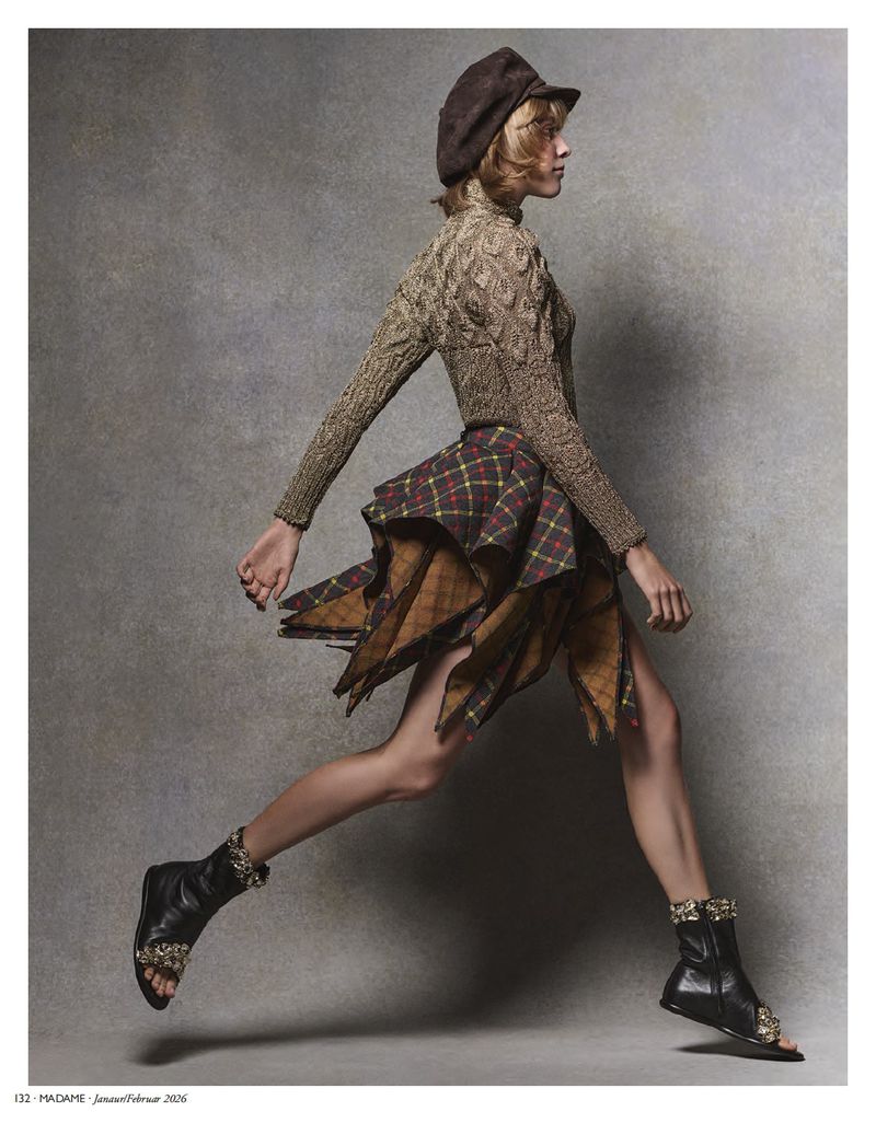 A stylish woman in a hat, sweater, and plaid skirt is captured in motion, leaping against a textured grey background, accessorized with unique boots.