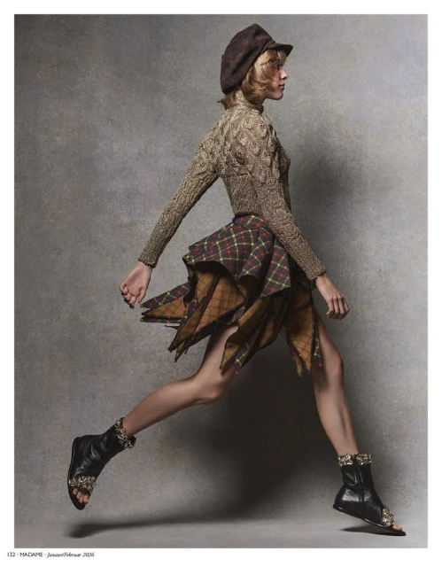 A stylish woman in a hat, sweater, and plaid skirt is captured in motion, leaping against a textured grey background, accessorized with unique boots.