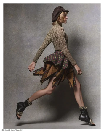 A stylish woman in a hat, sweater, and plaid skirt is captured in motion, leaping against a textured grey background, accessorized with unique boots.