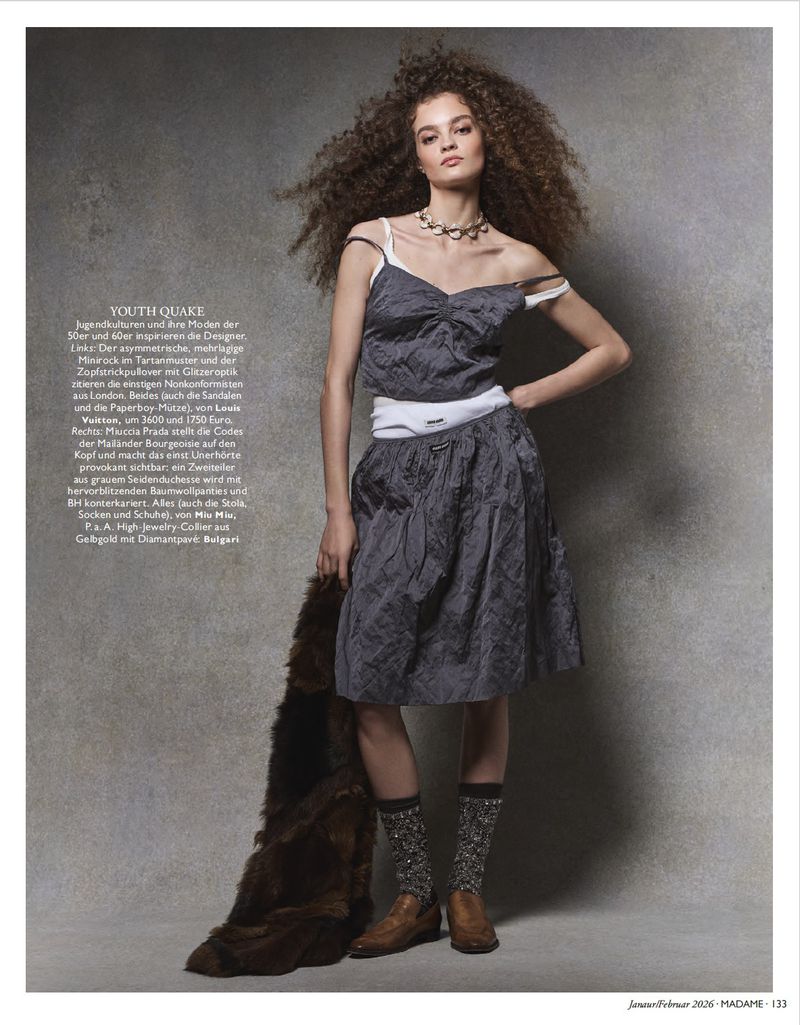 Fashion editorial shot of a young woman with curly brown hair in a gray dress, holding a fur shawl, posing in a studio with gray background.