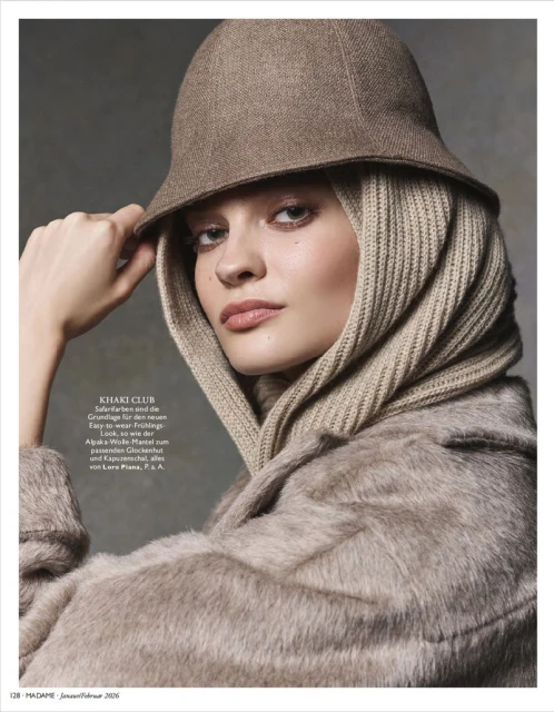 Close-up of a woman wearing a beige hat and hooded scarf, styled with a furry coat, looking directly at the camera in a studio setting.