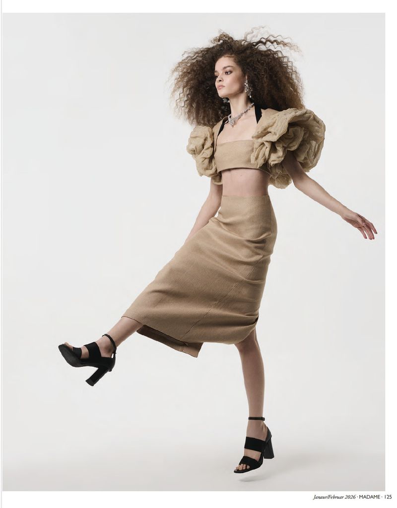 Fashion editorial shot of a woman in a beige crop top, skirt, and black high heels striking a pose in a studio setting.