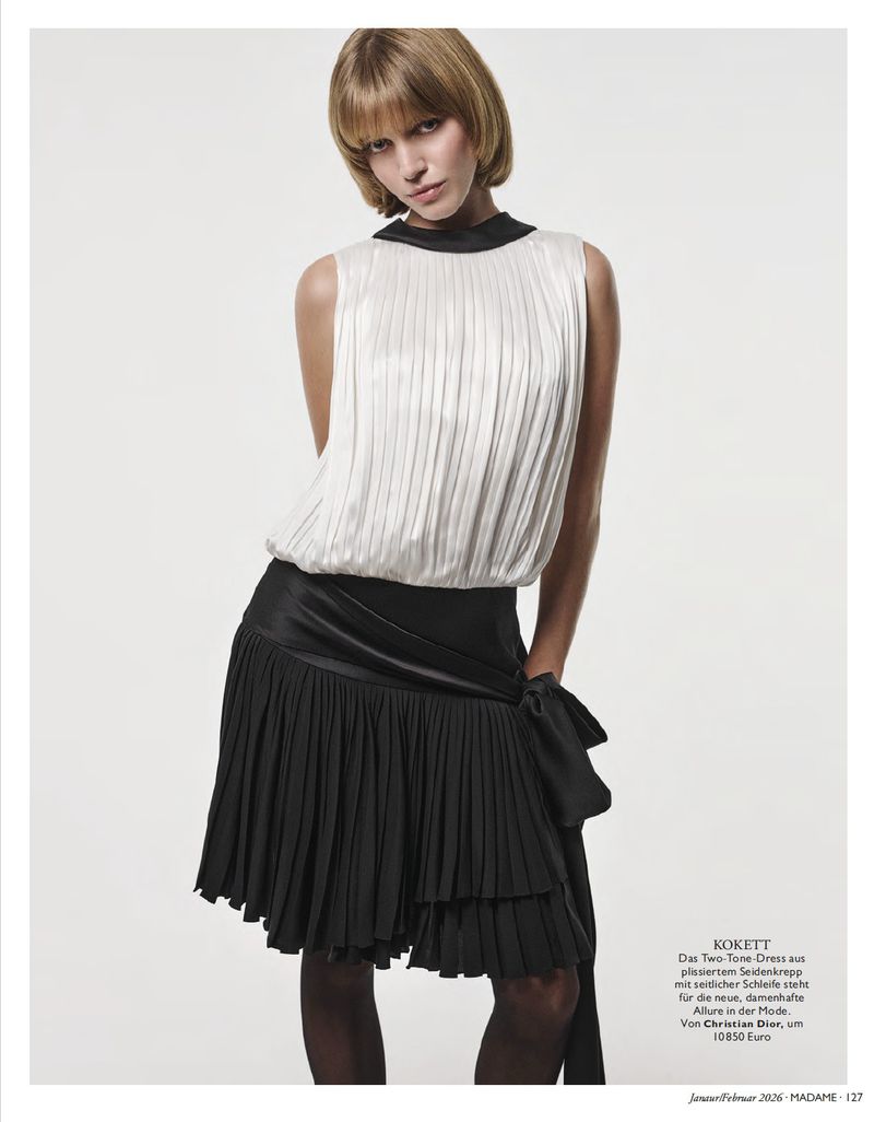 Blonde woman in a sleeveless white top and black pleated skirt poses against a neutral background in a fashion editorial shot.