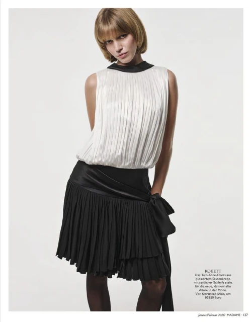 Blonde woman in a sleeveless white top and black pleated skirt poses against a neutral background in a fashion editorial shot.