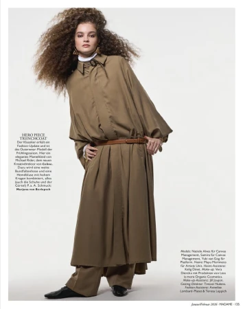 Model with voluminous curly hair wearing an oversized belted tan coat dress and wide trousers against a white studio background.