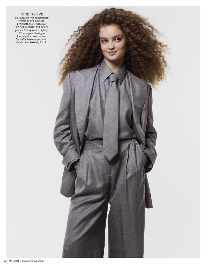 A woman with curly auburn hair poses in a grey power suit, matching tie, and collared shirt against a bright neutral background.