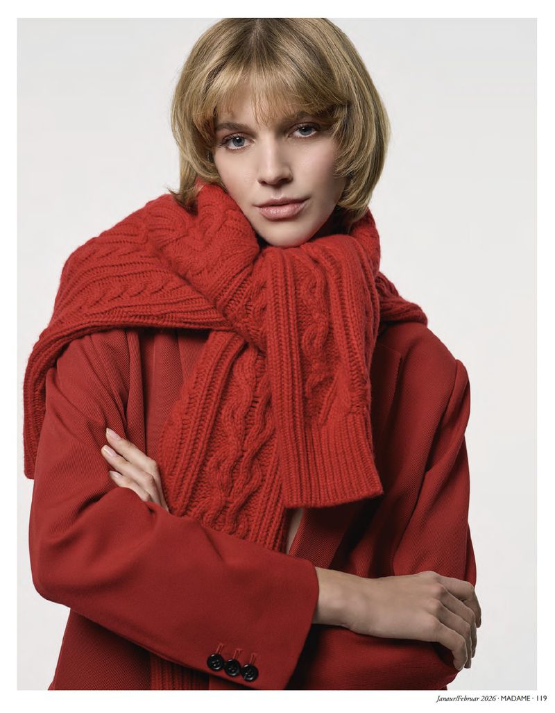 Portrait of a blonde woman wearing a red coat and knitted scarf, arms crossed, looking at the camera against a white background.