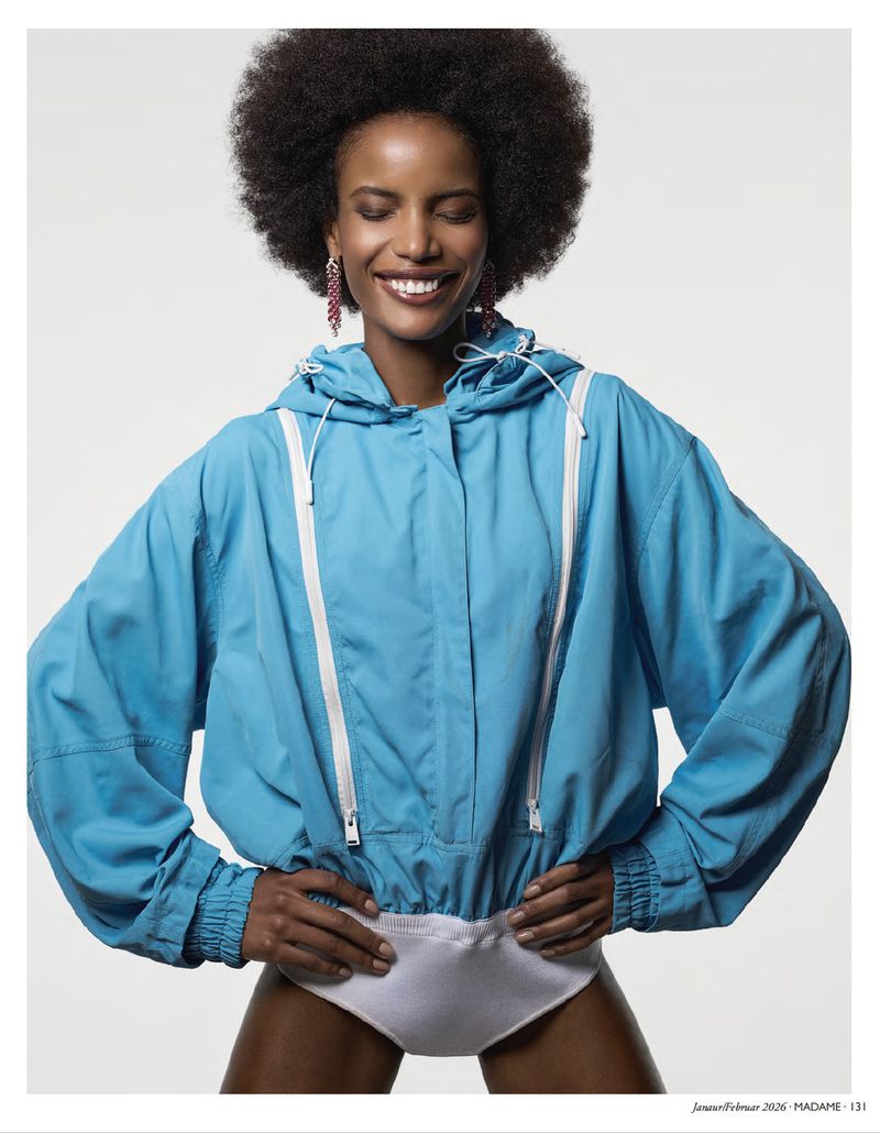 A smiling Black woman with an afro stands indoors wearing a blue hooded jacket and white bottoms, hands on hips, against a white backdrop.