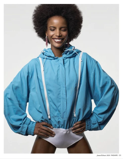 A smiling Black woman with an afro stands indoors wearing a blue hooded jacket and white bottoms, hands on hips, against a white backdrop.