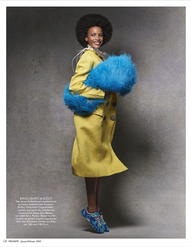 Model Natiele Alves smiling in a long yellow coat and oversized fluffy blue stole against a textured grey studio backdrop.