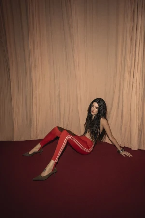 Brunette woman in red leggings and heels poses on a red carpet in front of a peach curtain backdrop in a fashion style shot.
