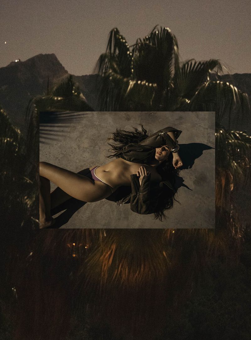 Adult female reclining in lingerie and jacket against a geometric shape, outdoor nighttime setting with palm trees and distant mountains.