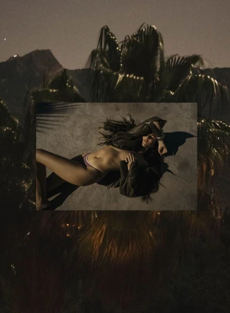 Adult female reclining in lingerie and jacket against a geometric shape, outdoor nighttime setting with palm trees and distant mountains.