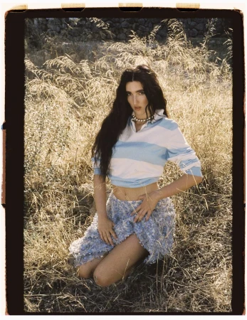 Brunette woman kneeling in sunlit field of tall grass, wearing a blue and white cropped polo and textured mini skirt, gazing at viewer.