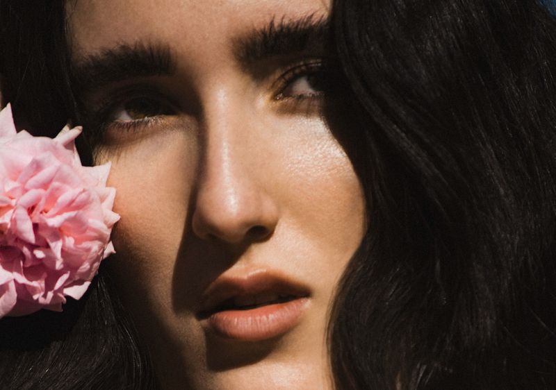Close-up portrait of a fair-skinned woman with dark hair and brows, with a large pink rose near her face in an outdoor setting.