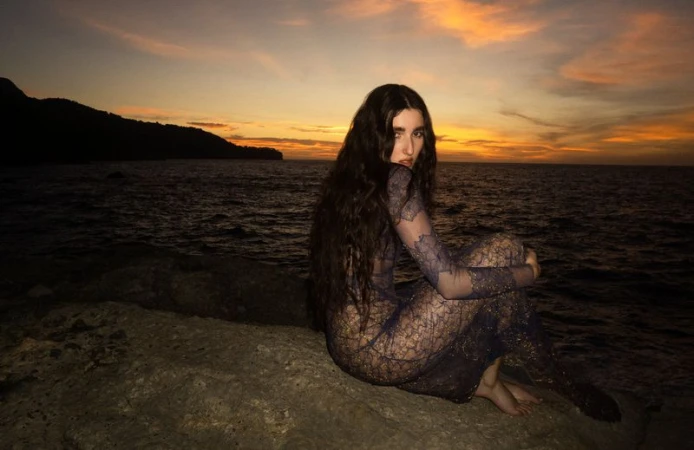A young woman with long dark hair sits on a rock outcrop at dusk near the sea, wearing a lace dress, with a silhouetted coastline.