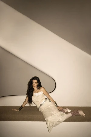 Adult female fashion model in a flowing white dress reclines on a brown bench in front of a white, geometric architectural wall. 