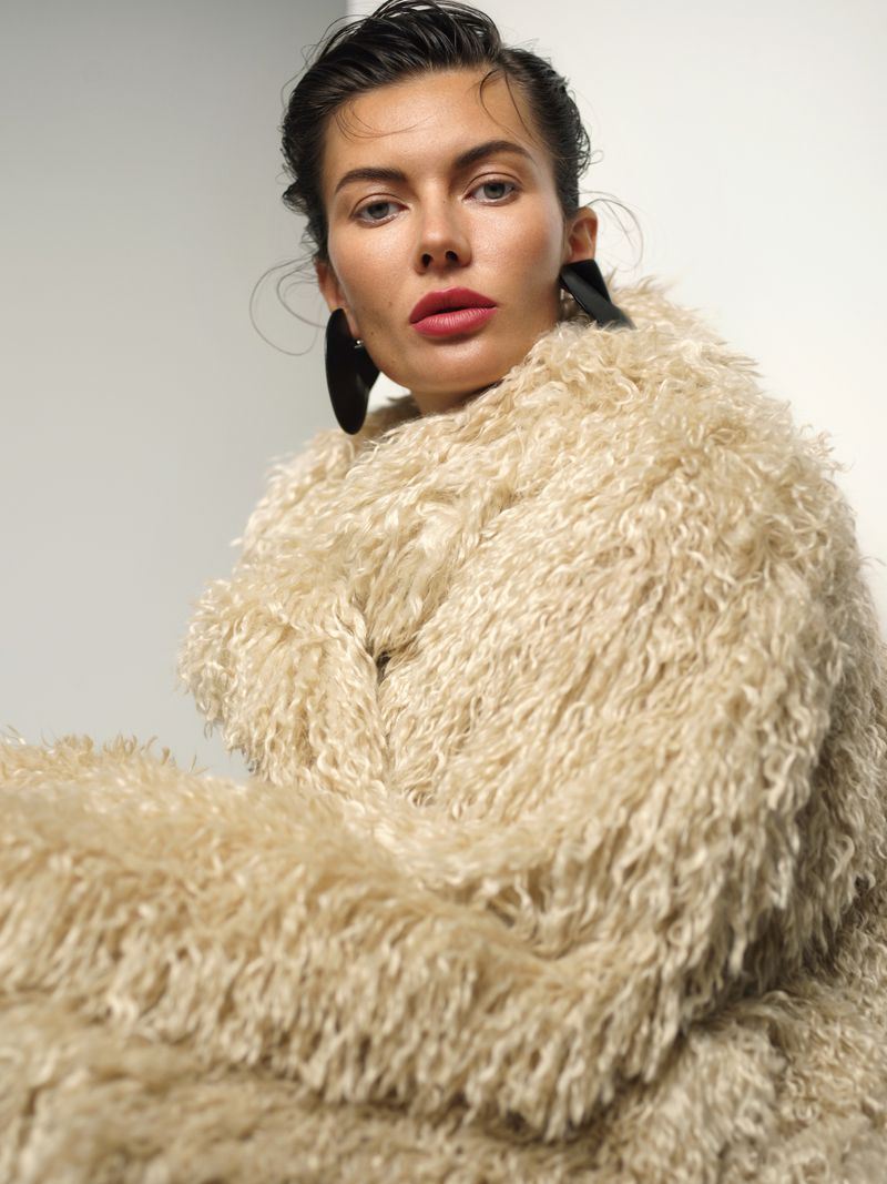 Woman wearing a textured cream shaggy coat and large black earrings against a plain white background
