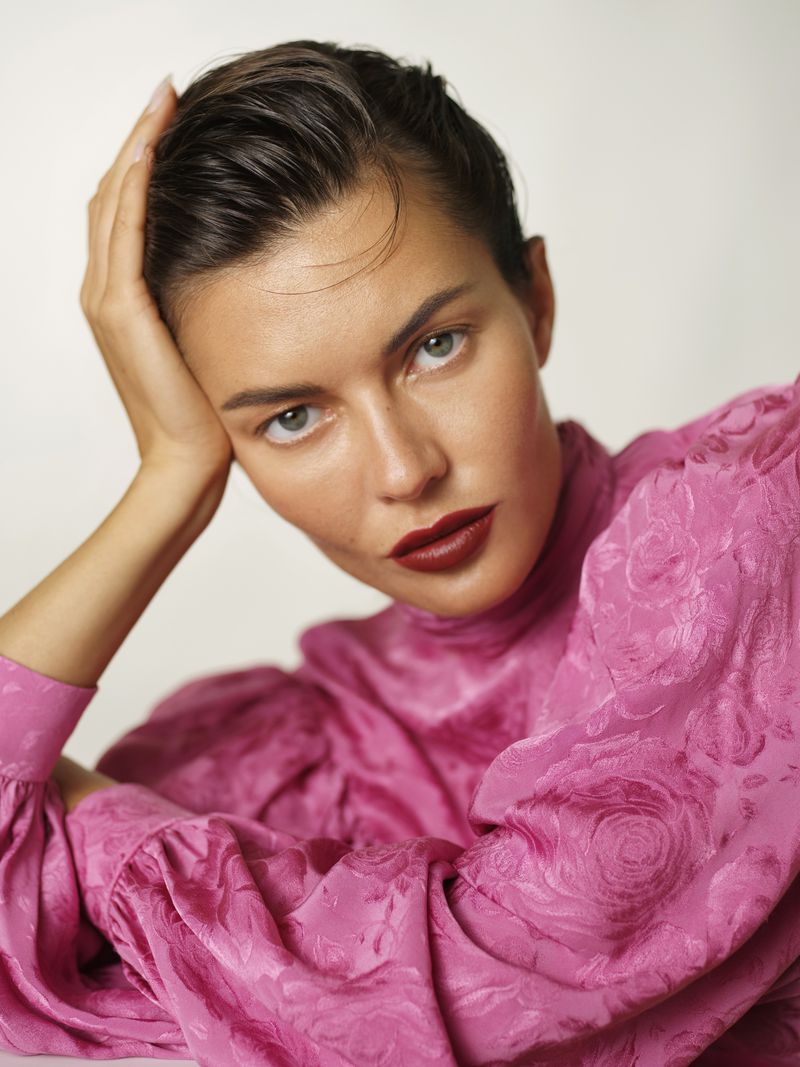 Close-up beauty portrait of a woman with slicked hair wearing bold dark red lipstick and a textured fuchsia pink puff-sleeve blouse.