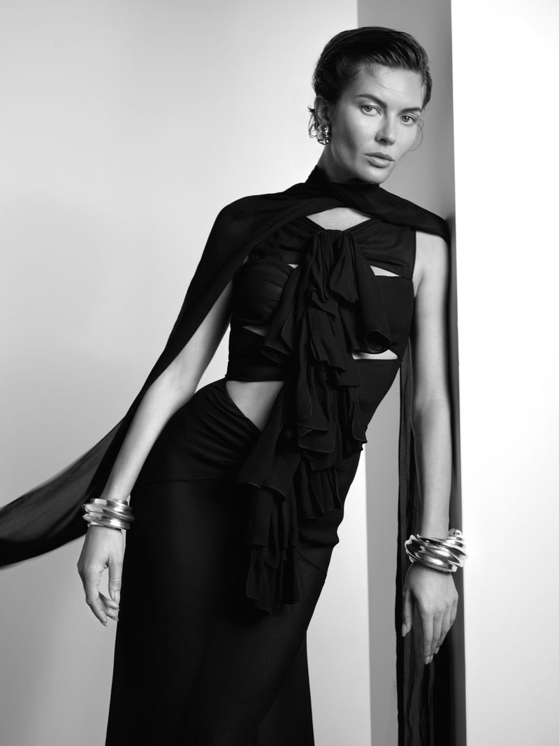 Black and white portrait of a woman wearing a stylish black dress with ruffles and silver bracelets leaning against a white wall.