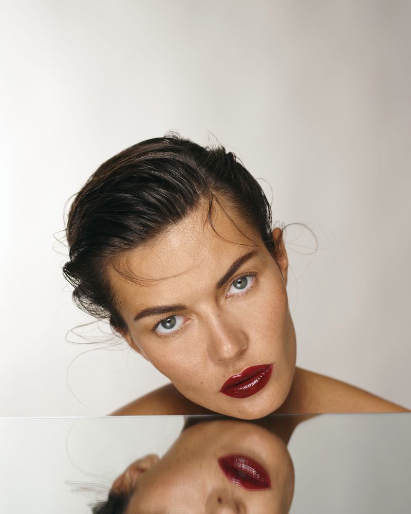 Close-up studio shot of a young adult woman with dark, slicked-back hair and red lips, reflected in a mirror on a clean background.
