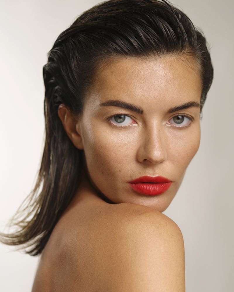 Close-up portrait of a woman with wet, slicked-back hair and bold red lipstick looking over her shoulder in a brightly lit photo studio.